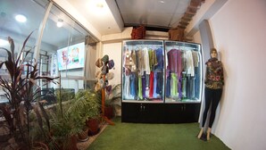 Ein Zimmer mit einer in schwarze Hose und ein gemustertes Oberteil gekleideten Mannequin, neben einem Kleiderständer mit verschiedenen Kleidungsstücken. Auf einem Regal stehen Blumentöpfe und ein Fernseher zeigt einen Zeichentrickfilm.
