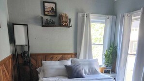 A bedroom with a bed, a bedside table with a lamp, a fan, and a window with curtains.