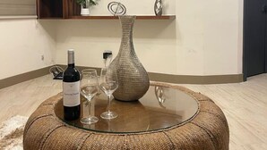 Un salon avec une table basse ronde en verre, une bouteille de vin et deux verres posés dessus. Un grand téléviseur à écran plat est fixé au mur, au-dessus d’une étagère en bois sur laquelle se trouve un pot de fleurs.