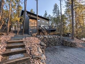 Une maison moderne à deux étages, dotée d’un mur de soutènement en pierre et d’un escalier en bois menant à une terrasse.