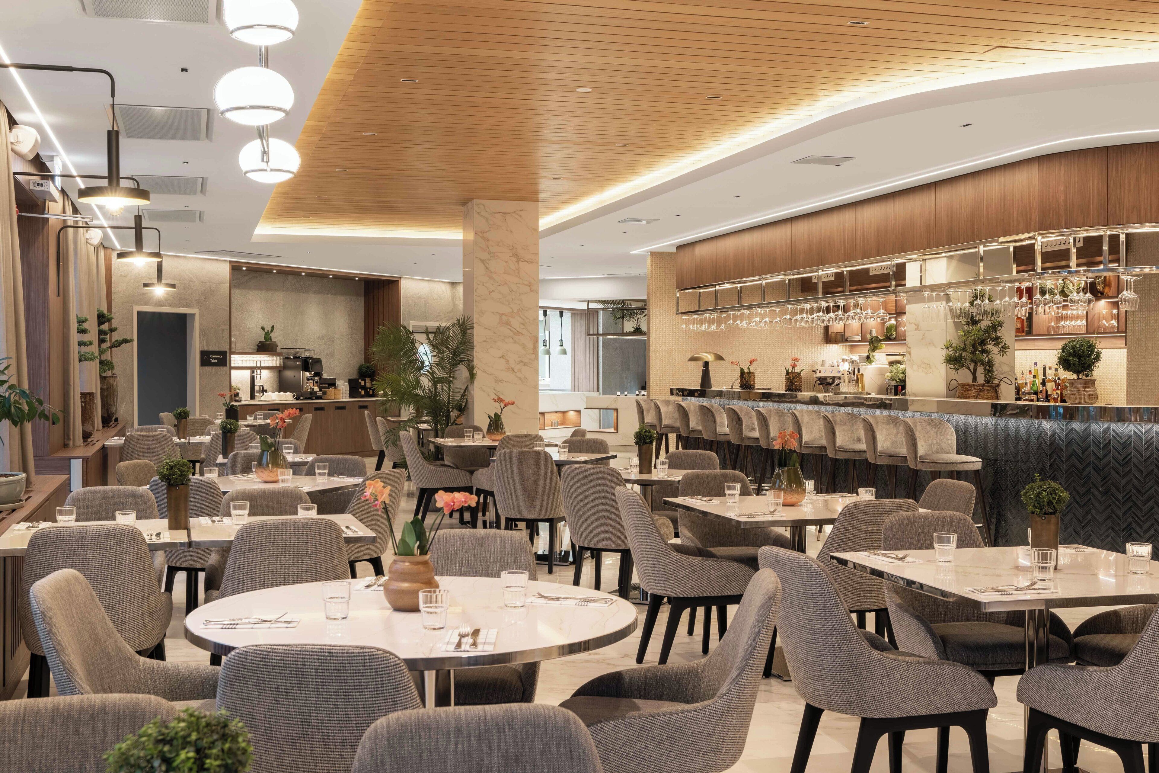 Un restaurant moderne avec des tables rondes et des chaises grises, un bar avec des verres à vin et une cuisine visible au fond de la salle.