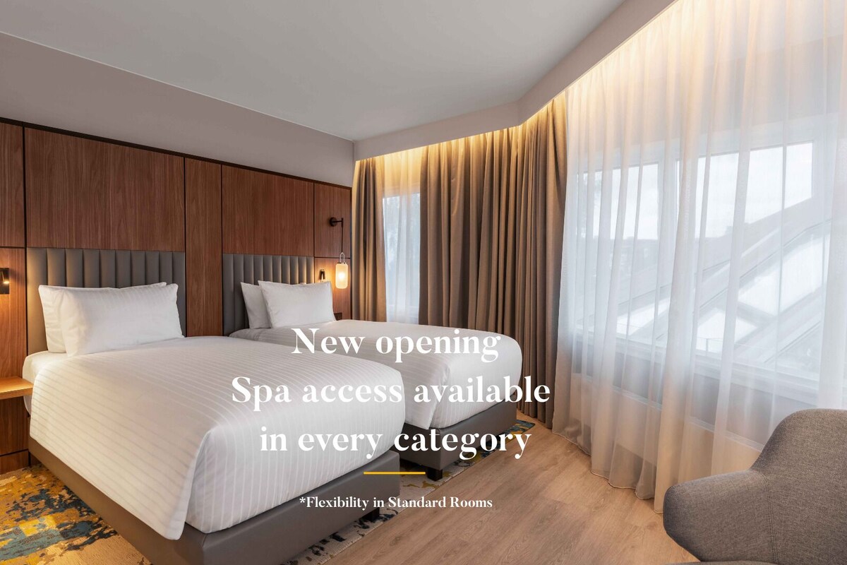 A modern hotel room with a large bed, wooden headboard, and a view of the city through sheer curtains.