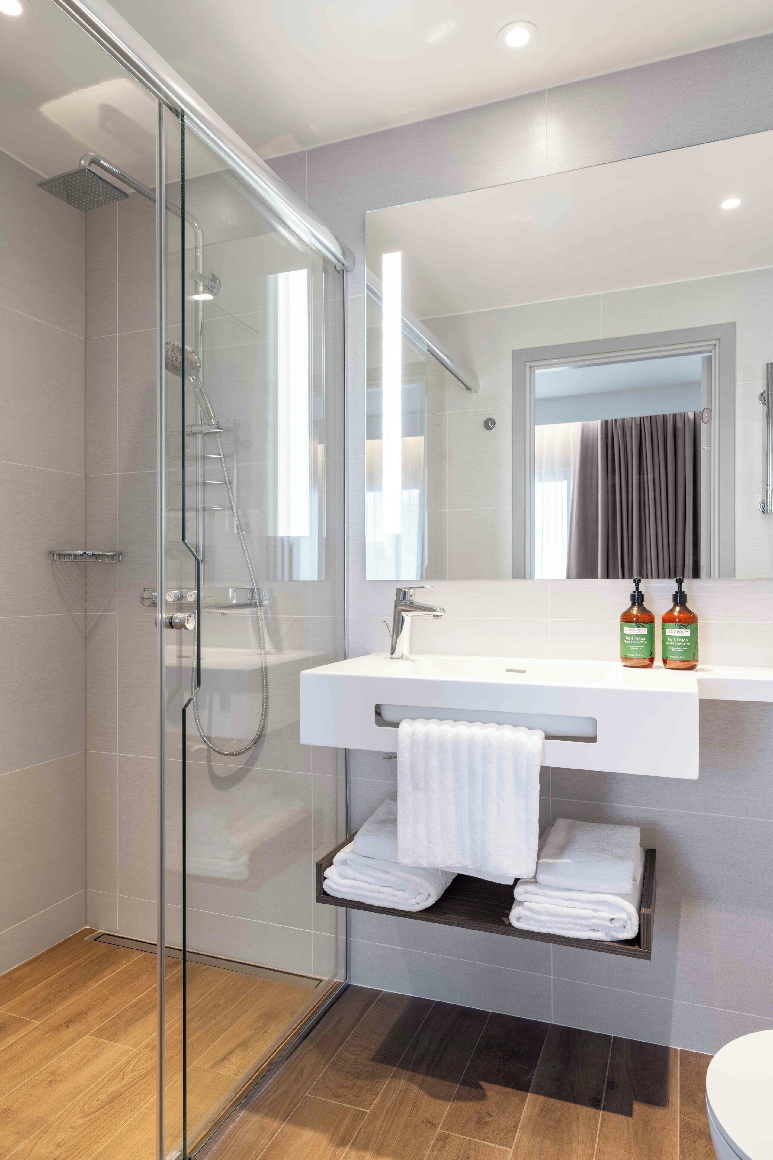 A modern bathroom with a glass-enclosed shower, a white sink with a mirror above, and neatly folded towels on a shelf.