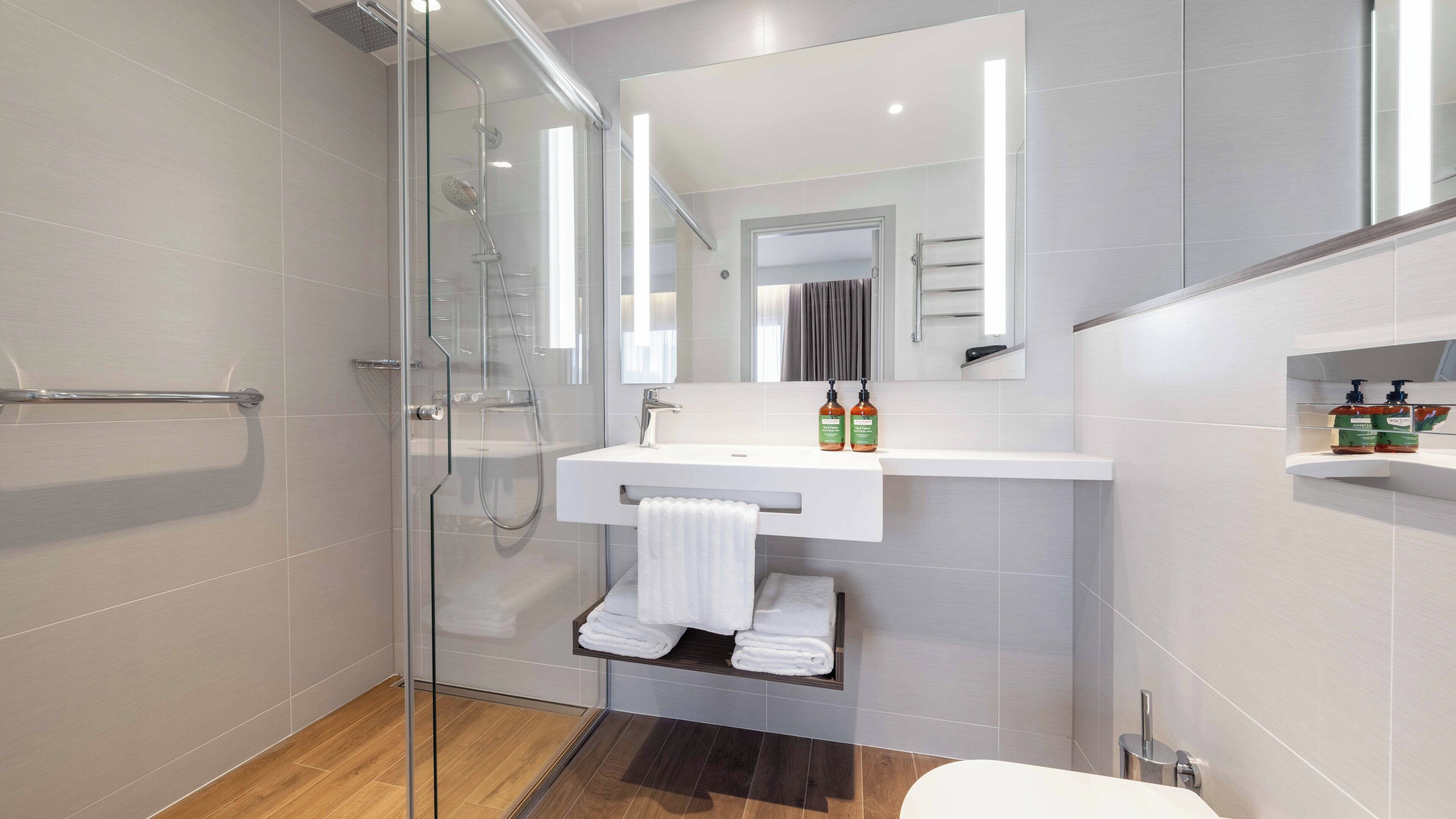 A modern bathroom with a glass shower enclosure, a white sink, and a mirror above it. There are two bottles of shampoo on the sink.