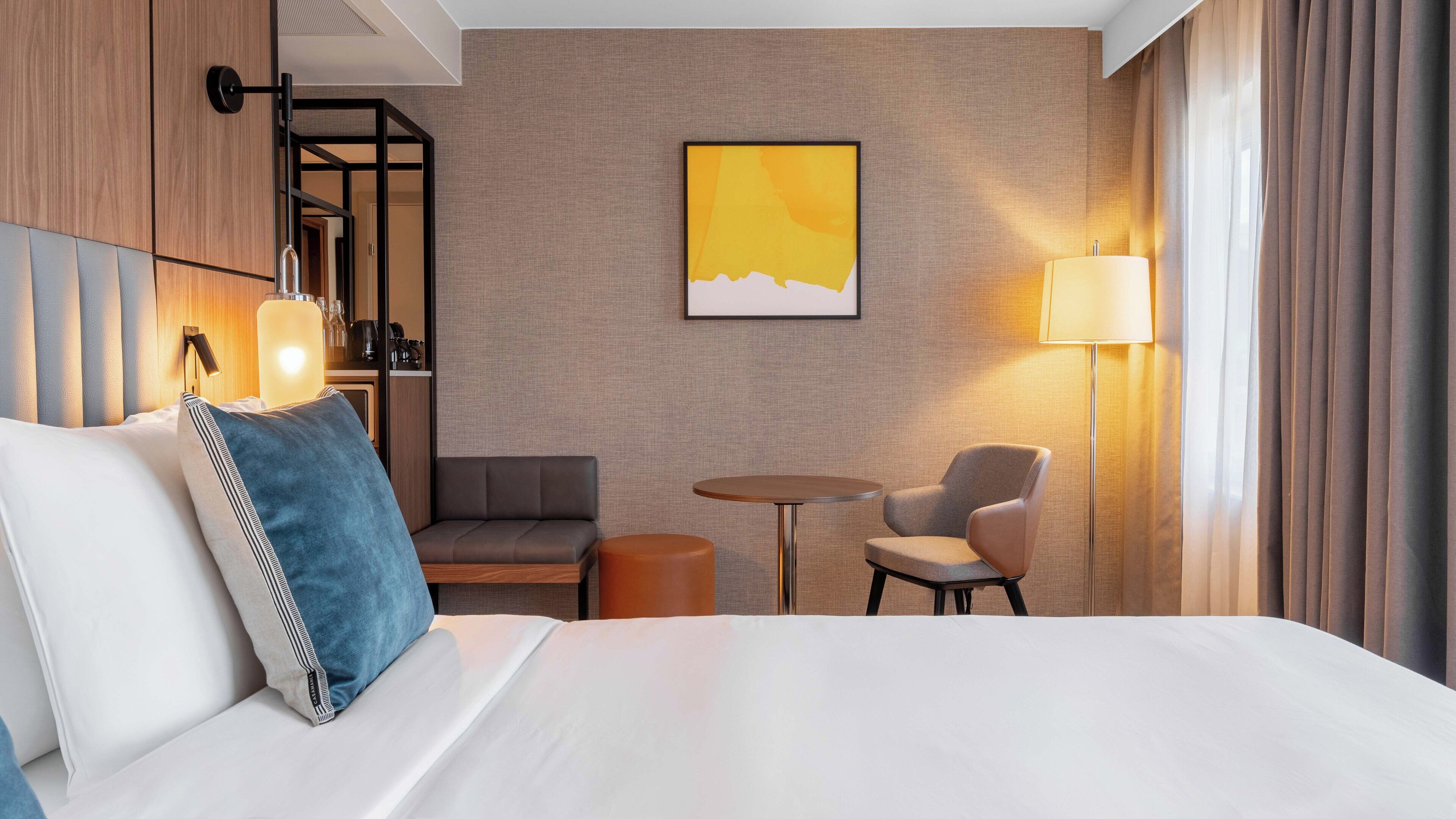 A hotel room with a bed, a seating area with a table and chairs, a lamp, and a wall-mounted artwork.