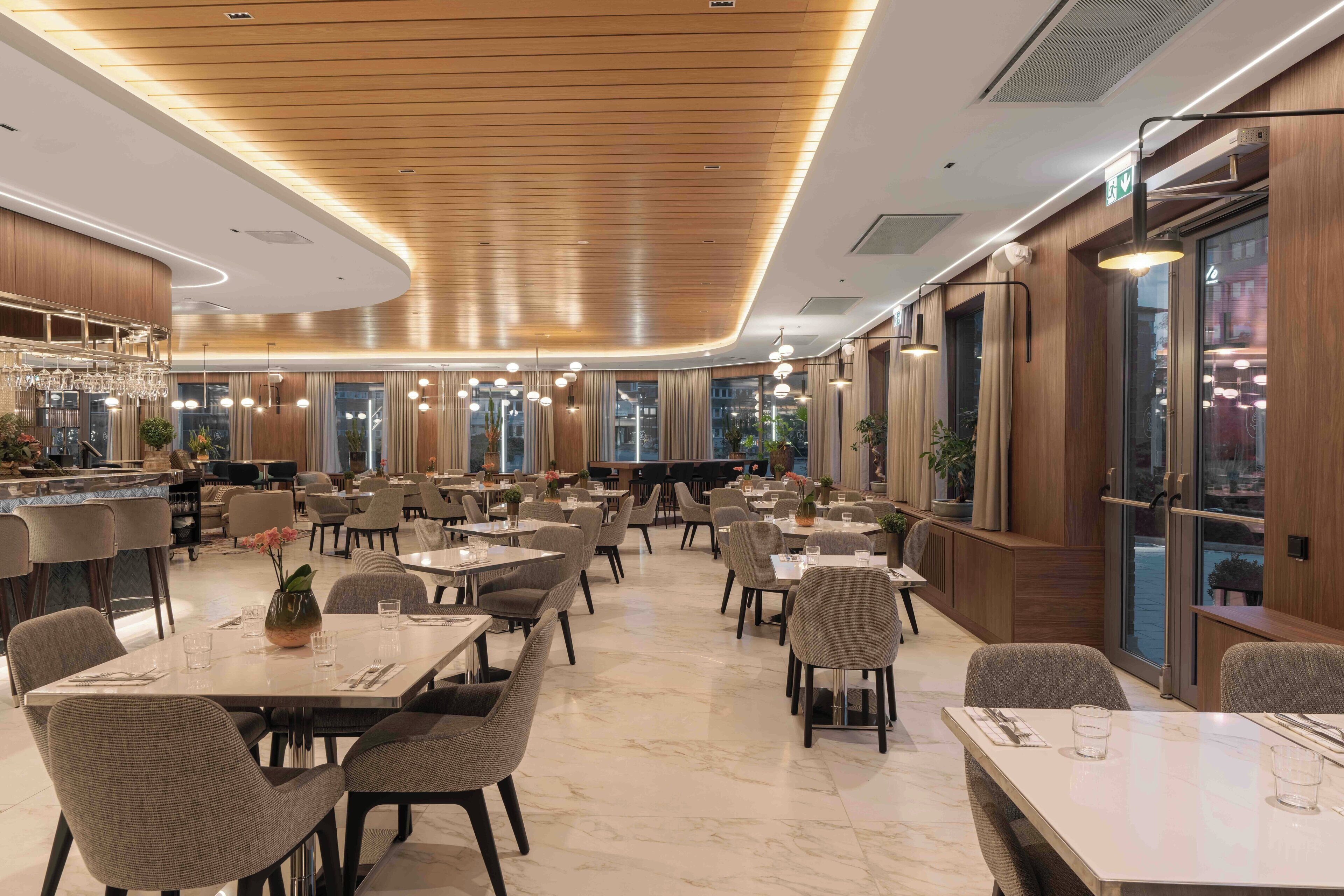 A spacious hotel dining area with multiple tables and chairs, a bar area with a counter and hanging lights, and large windows providing views of the city.