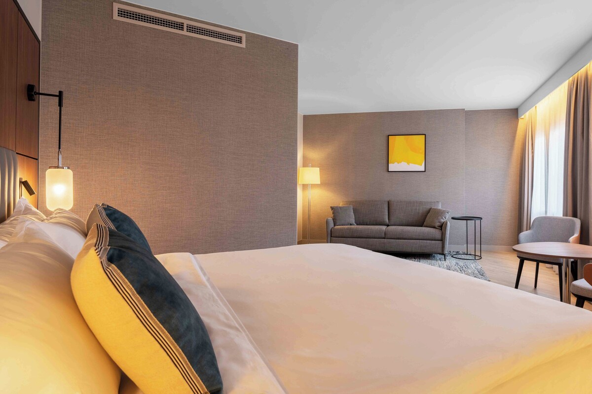 A hotel room with a large bed, a sofa, a small table, and a chair.