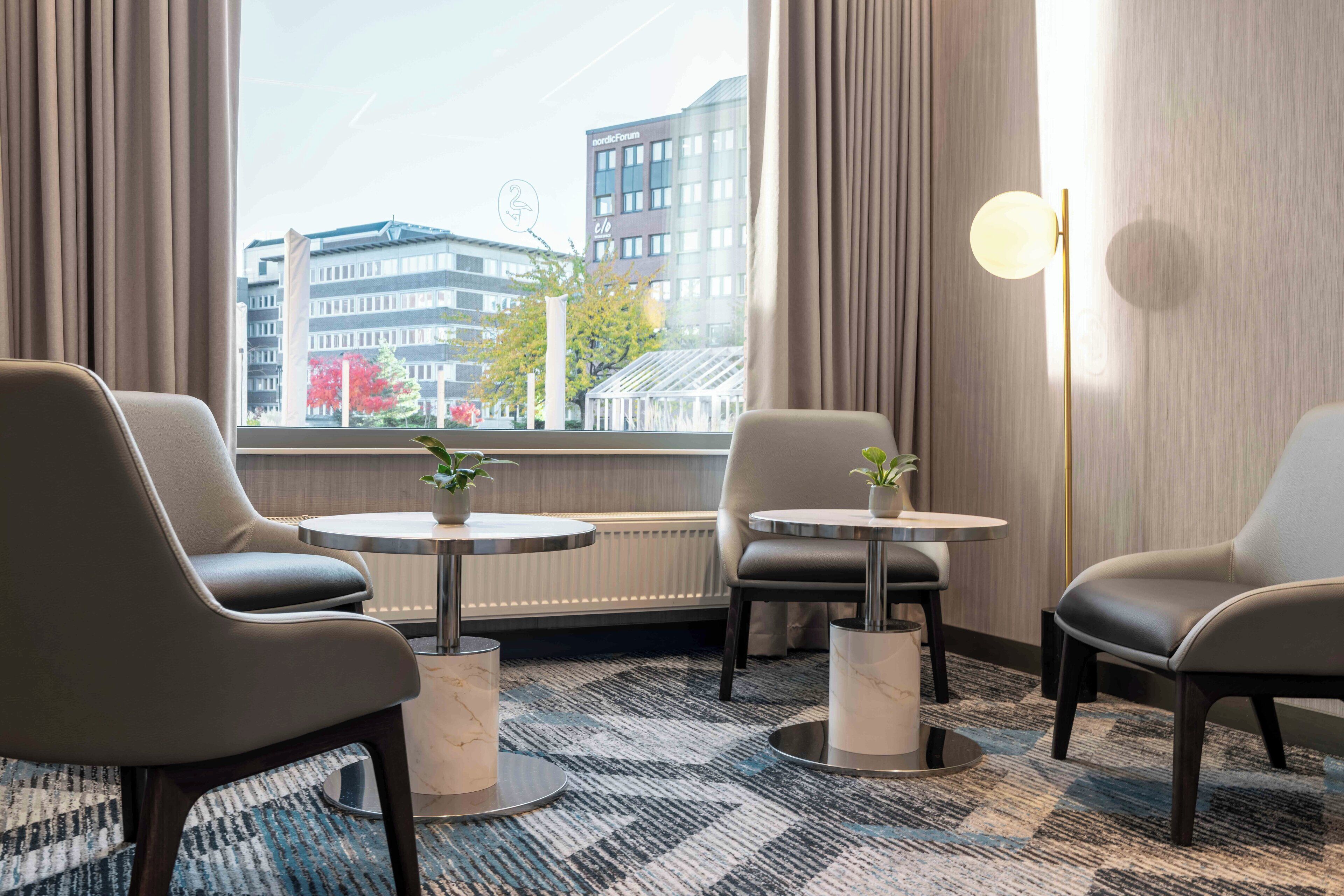 A modern hotel room with two leather chairs, a small table with a plant, and a floor lamp. A large window offers a view of city buildings.