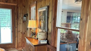 A wooden interior with a wooden ceiling, a wall-mounted clock, a framed painting, a lamp, and a window with a view of the outdoors.