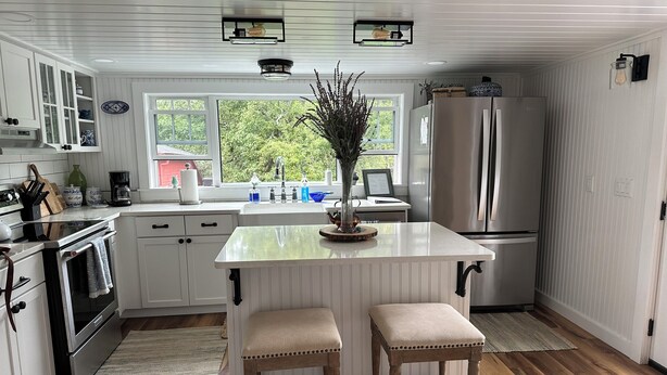 A kitchen with white cabinetry, a central island, two barstools, a stainless steel refrigerator, and a window with a view of greenery.