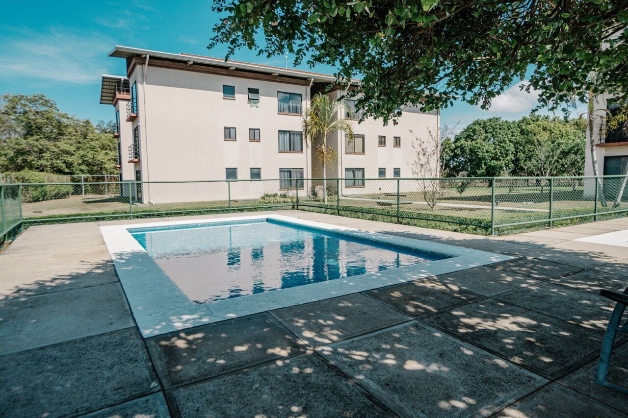 A swimming pool with a tiled deck, surrounded by a fence and trees.