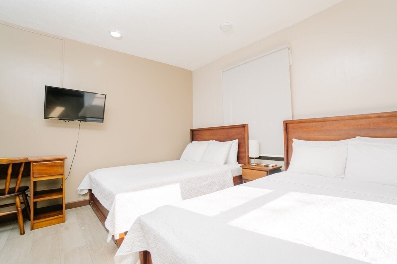 A hotel room with two single beds, a TV mounted on the wall, a wooden nightstand, and a desk with drawers.