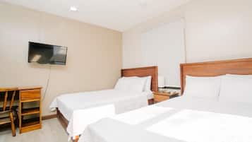 A hotel room with two single beds, a TV mounted on the wall, a wooden nightstand, and a desk with drawers.