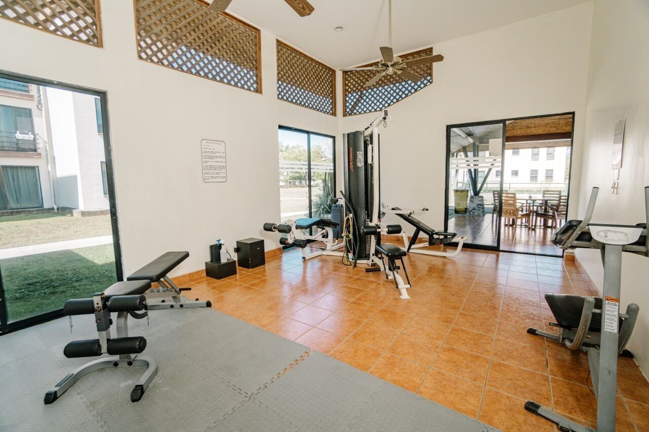 Ein Fitnessraum mit Trainingsgeräten, großen Fenstern und einem Deckenventilator.
