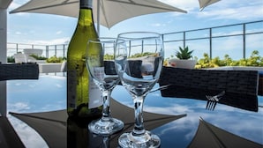 A table with a bottle of wine and two glasses, set outdoors with a clear sky and plants in the background.