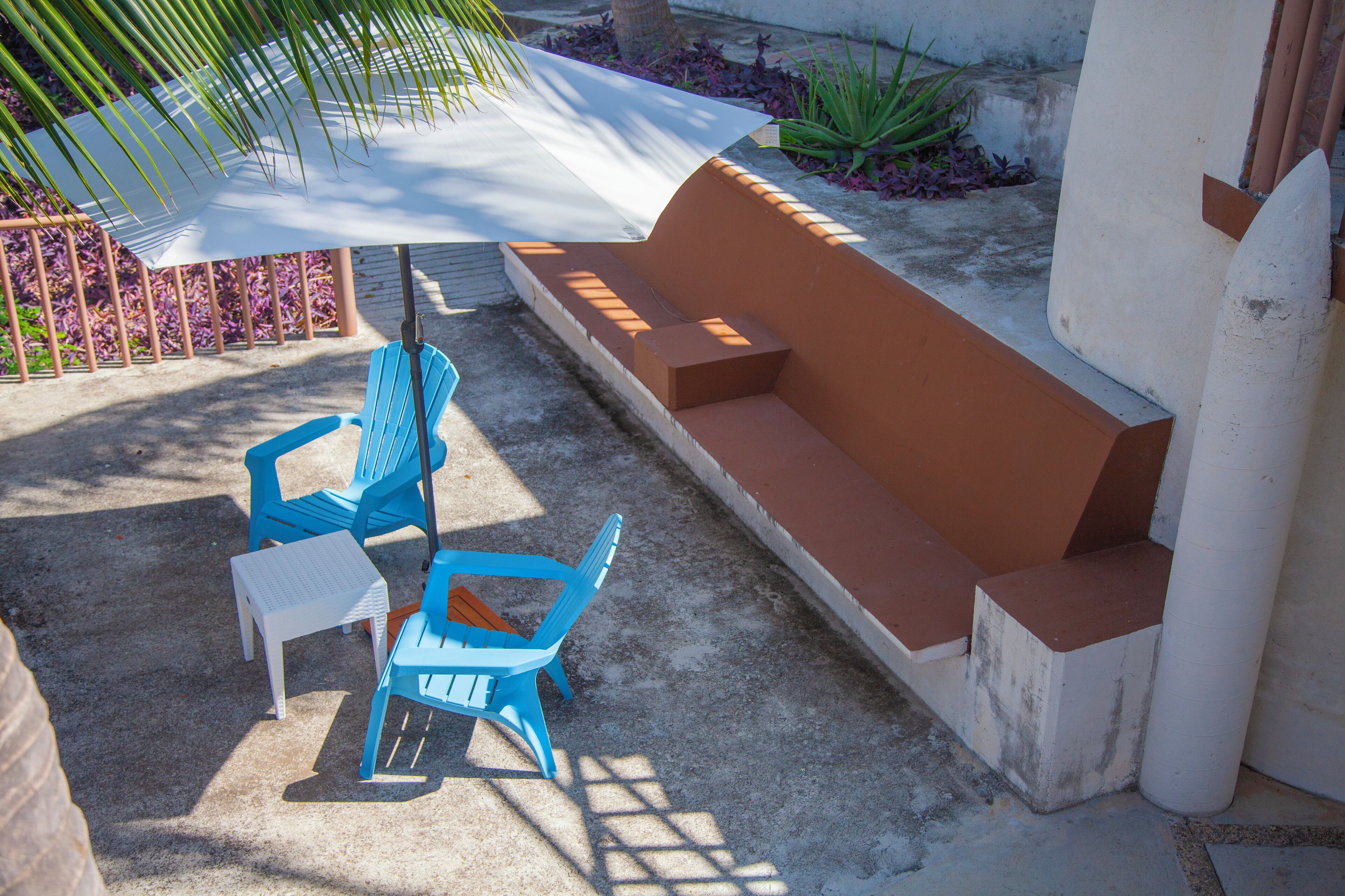 A patio area with a white umbrella, blue chairs, and a brown bench.