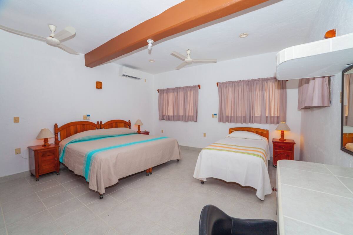 Une chambre d’hôtel avec deux lits, des têtes de lit en bois, des tables de chevet avec des lampes, un ventilateur de plafond et un grand miroir au mur.