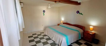 Un dormitorio con piso a cuadros, una cama con colcha azul y blanca, dos adornos florales en la pared y un ventilador de techo.