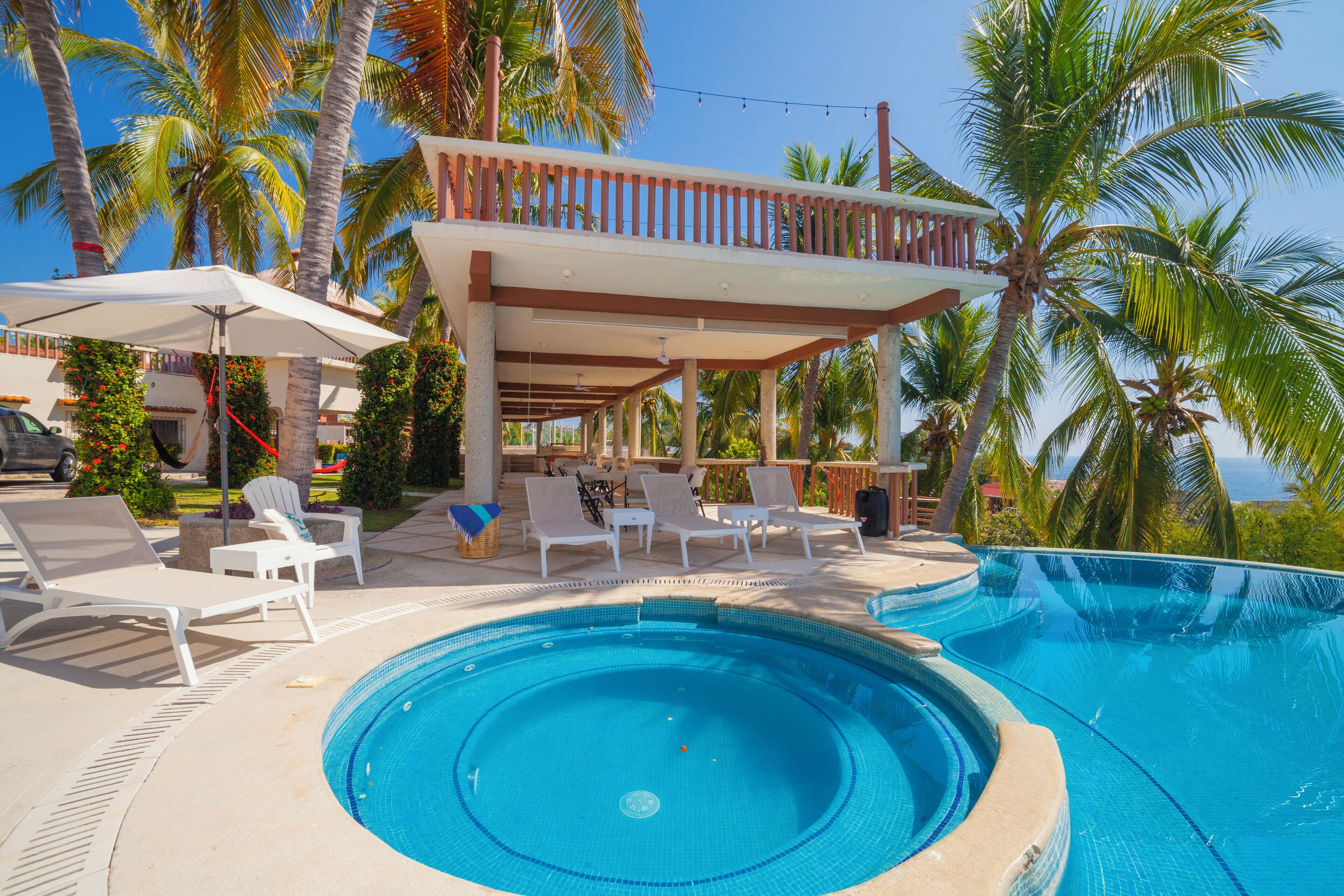 Un área de piscina con sillas de descanso, una zona de estar cubierta y palmeras.