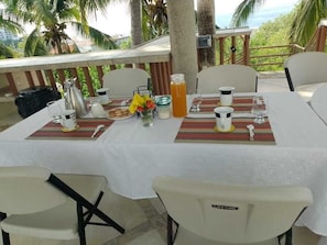 A table set for breakfast with coffee, tea, and juice, surrounded by chairs with a view of the ocean and palm trees. - Hotel Casa Rush (San Pedro Pochutla)