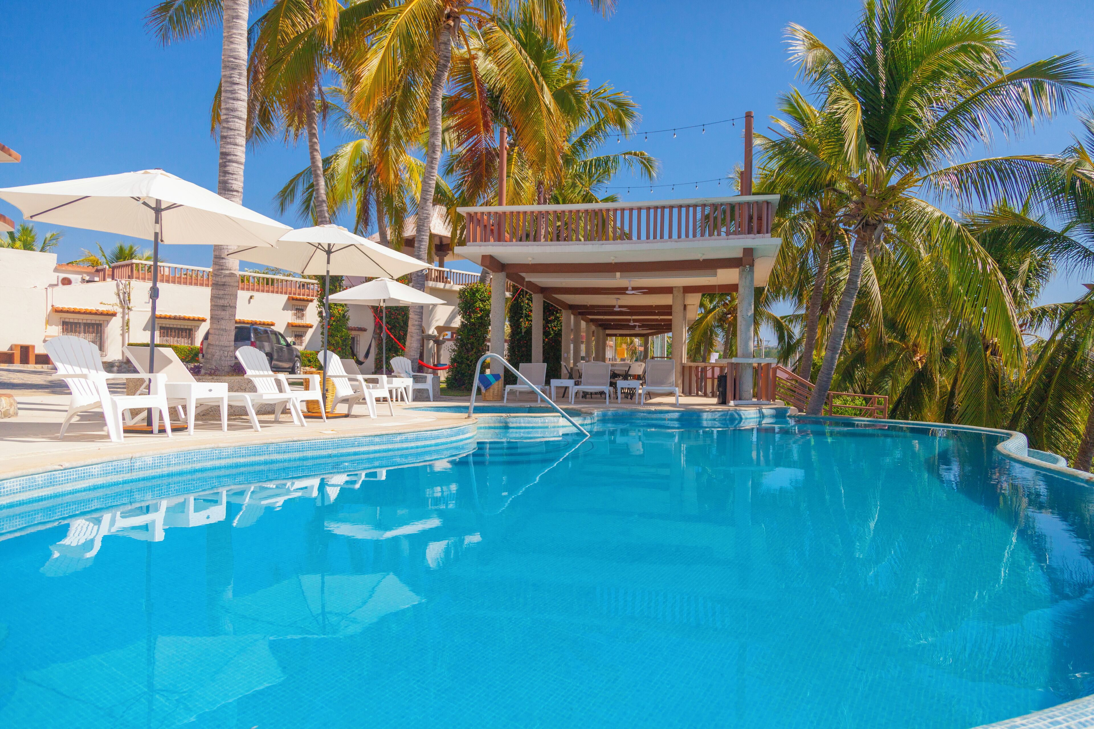 A swimming pool with lounge chairs and umbrellas, surrounded by palm trees and a building with a balcony.