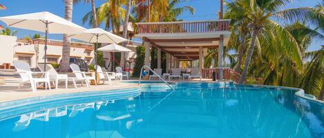 A swimming pool with lounge chairs and umbrellas, surrounded by palm trees and a building with a balcony.