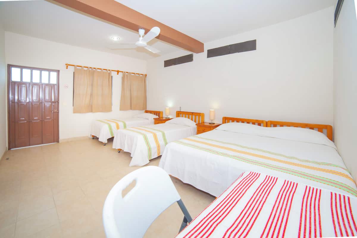 Une chambre d’hôtel avec trois lits simples, un ventilateur de plafond, une porte en bois et une chaise blanche.
