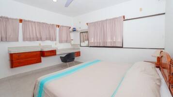 A bedroom with a bed, a desk, a chair, a vanity with a mirror, and a ceiling fan.