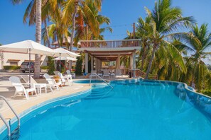 A swimming pool with lounge chairs and umbrellas, surrounded by palm trees and a covered area with a railing. - Hotel Casa Rush (San Pedro Pochutla)