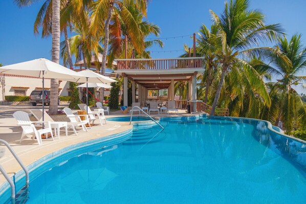 Une piscine entourée de palmiers, avec des chaises longues et des parasols, ainsi qu’un espace couvert doté d’une rambarde.