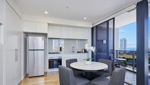 A modern kitchen with white cabinets, a stainless steel refrigerator, and an oven. There is a dining area with a round table and grey chairs.