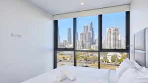 A modern hotel room with a large bed, white linens, and a city view through floor-to-ceiling windows.