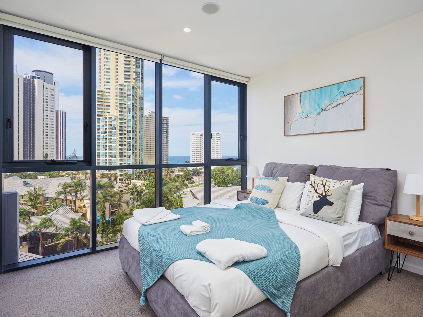 A bedroom with a large window, a bed with a teal blanket, and a city view.