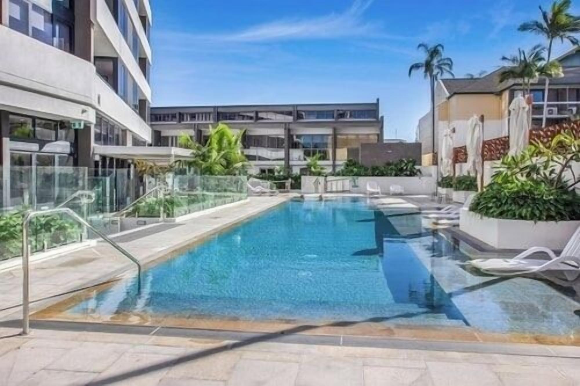 A rectangular pool with clear blue water, surrounded by a glass barrier, and adjacent to a modern building with balconies.