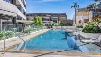 A rectangular pool with clear blue water, surrounded by a glass barrier, and adjacent to a modern building with balconies.