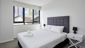 Una habitación de hotel moderna con una cama grande, un cabecero tapizado en gris, ropa de cama blanca, un ventanal con vistas a la ciudad y una mesita de noche con una lámpara.
