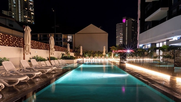 Una piscina nocturna con sillones de playa y sombrillas, rodeada de edificios.