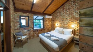 A bedroom with a brick wall, wooden ceiling beams, a bed with white linens, a wicker chair, and a window with a view of greenery.