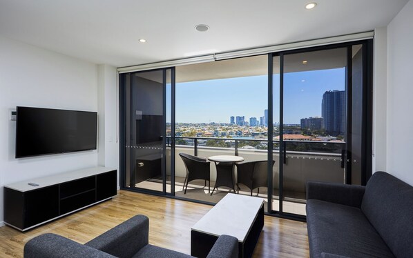 A modern living room with a flat-screen TV, a sofa, a coffee table, and a balcony with a city view.