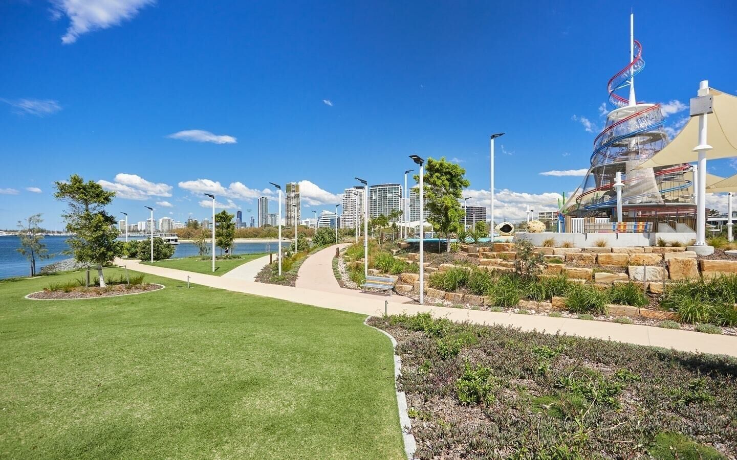 A waterfront park with pathways, greenery, and modern buildings in the background.