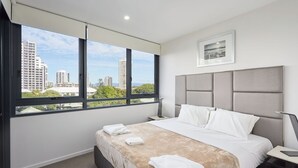 Moderni hotellihuone, jossa on suuri sänky, yöpöytä, pieni sivupöytä, ikkuna, josta on näkymä kaupunkiin, ja seinällä oleva kehystetty taulu.