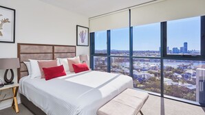 Ein modernes Hotelzimmer mit einem großen Bett, zwei Nachttischlampen, einer Bank und Blick auf die Stadt durch raumhohe Fenster.