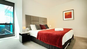 A modern hotel room with a large bed, a nightstand, a window with a city view, and a framed picture on the wall.