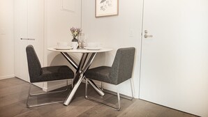 A dining area with a white table and two grey upholstered chairs, a vase with flowers, and a framed picture on the wall.
