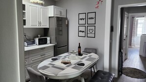 Ett modernt kök med vita skåp, en inbyggd mikrovågsugn och ett kylskåp. Det finns ett matbord med en rutig duk och en vinflaska.
