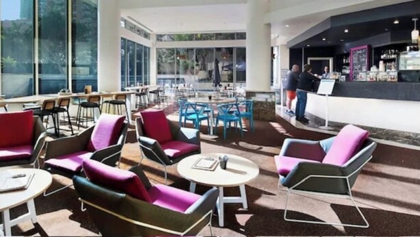 Un área de estar moderna con mesas y sillas, grandes ventanales con vistas a la ciudad y una barra de bar al fondo.
