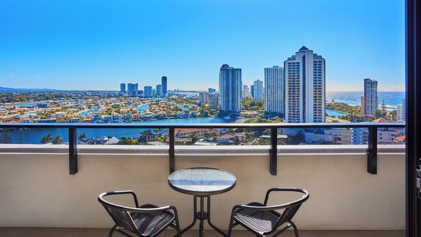 Un balcon avec deux chaises et une petite table, donnant sur un paysage urbain avec de hauts immeubles et une étendue d’eau.