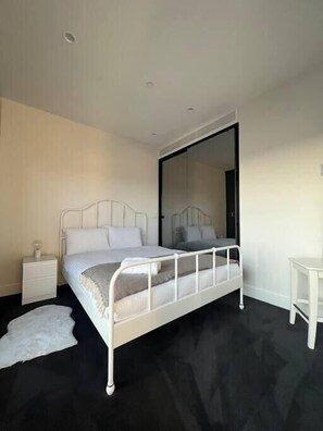 Un dormitorio con estructura de cama de metal blanco, una mesita de noche, una pequeña mesa y un espejo grande.