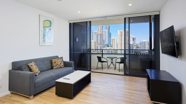 A modern living room with a grey sofa, a coffee table, and a TV. There is a balcony with a city view.