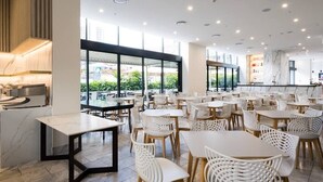 Un restaurant moderne avec des chaises et des tables blanches, de grandes fenêtres et une vue sur la verdure environnante.